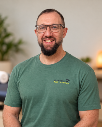 A smiling man with a beard and glasses wears a green “About Us” t-shirt featuring the text “Alternative Myotherapy.”